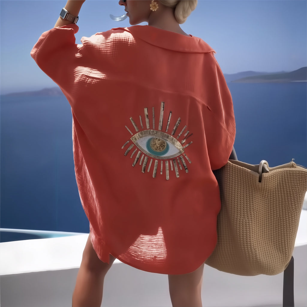 Woman wearing coral bikini with embroidered eye motif, standing by Mediterranean sea with beach bag