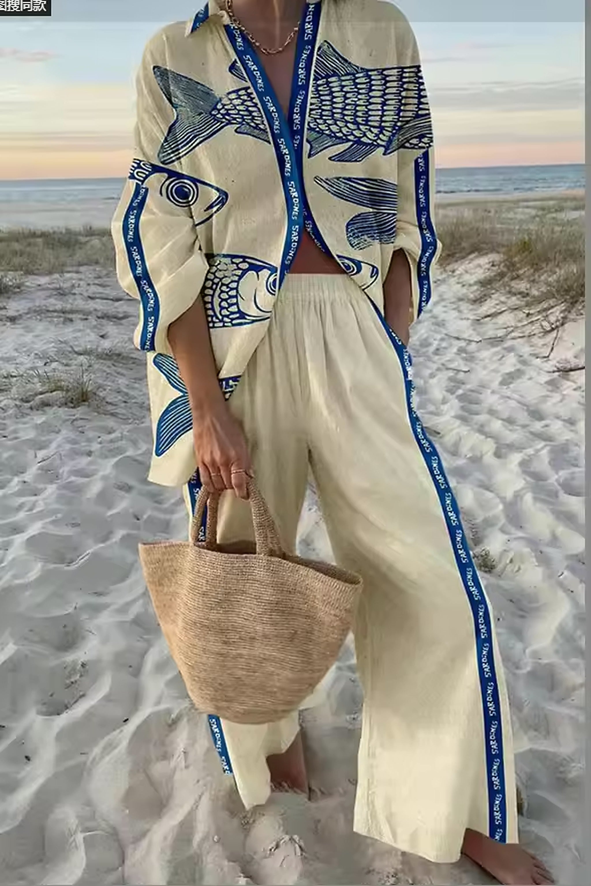 Woman wearing cream bikini top with navy fish print shirt on sandy beach at sunset, holding woven tote bag.