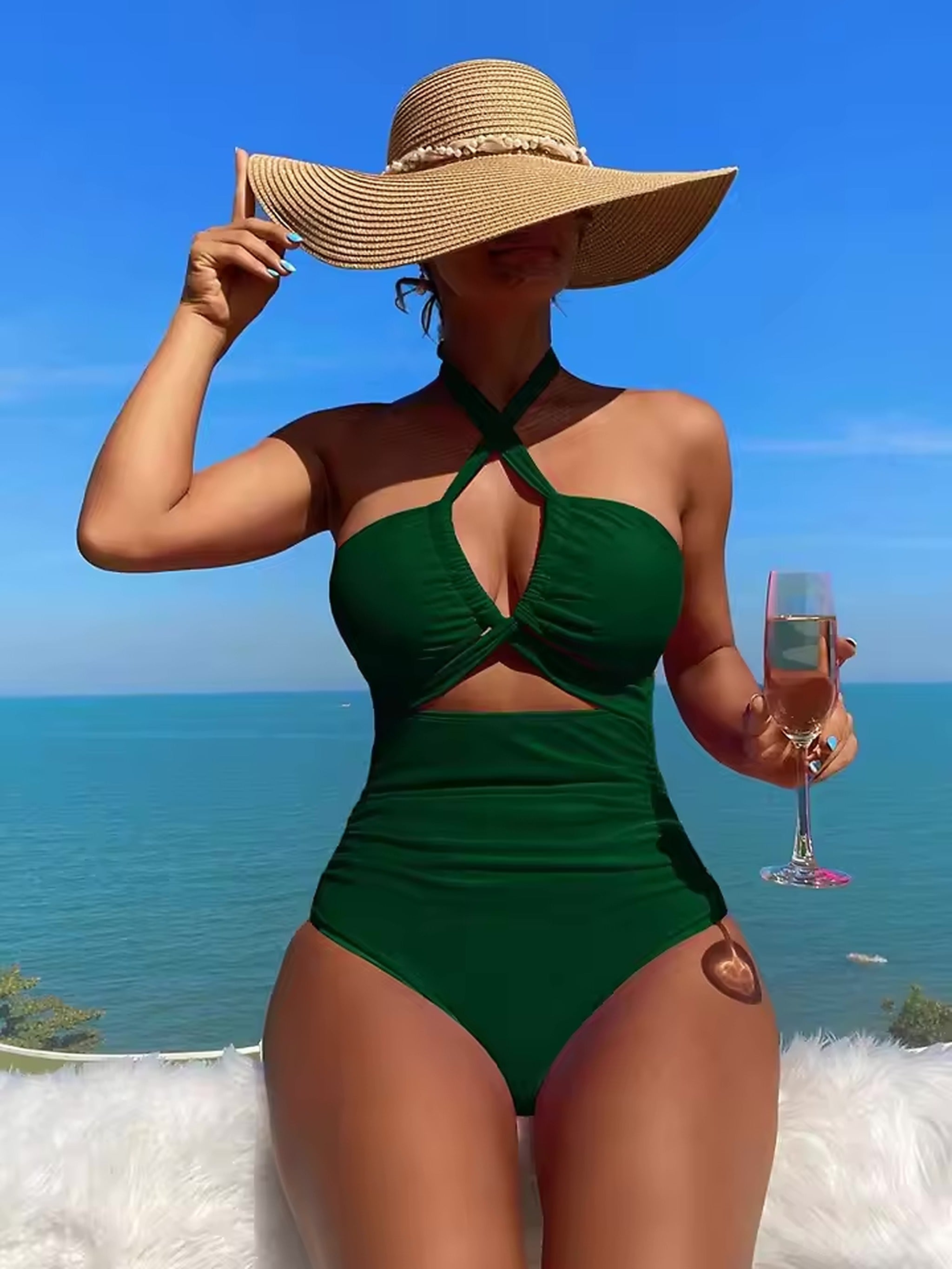 Woman wearing emerald green halter bikini with cutout detail, wide-brimmed straw hat, holding champagne glass by the sea