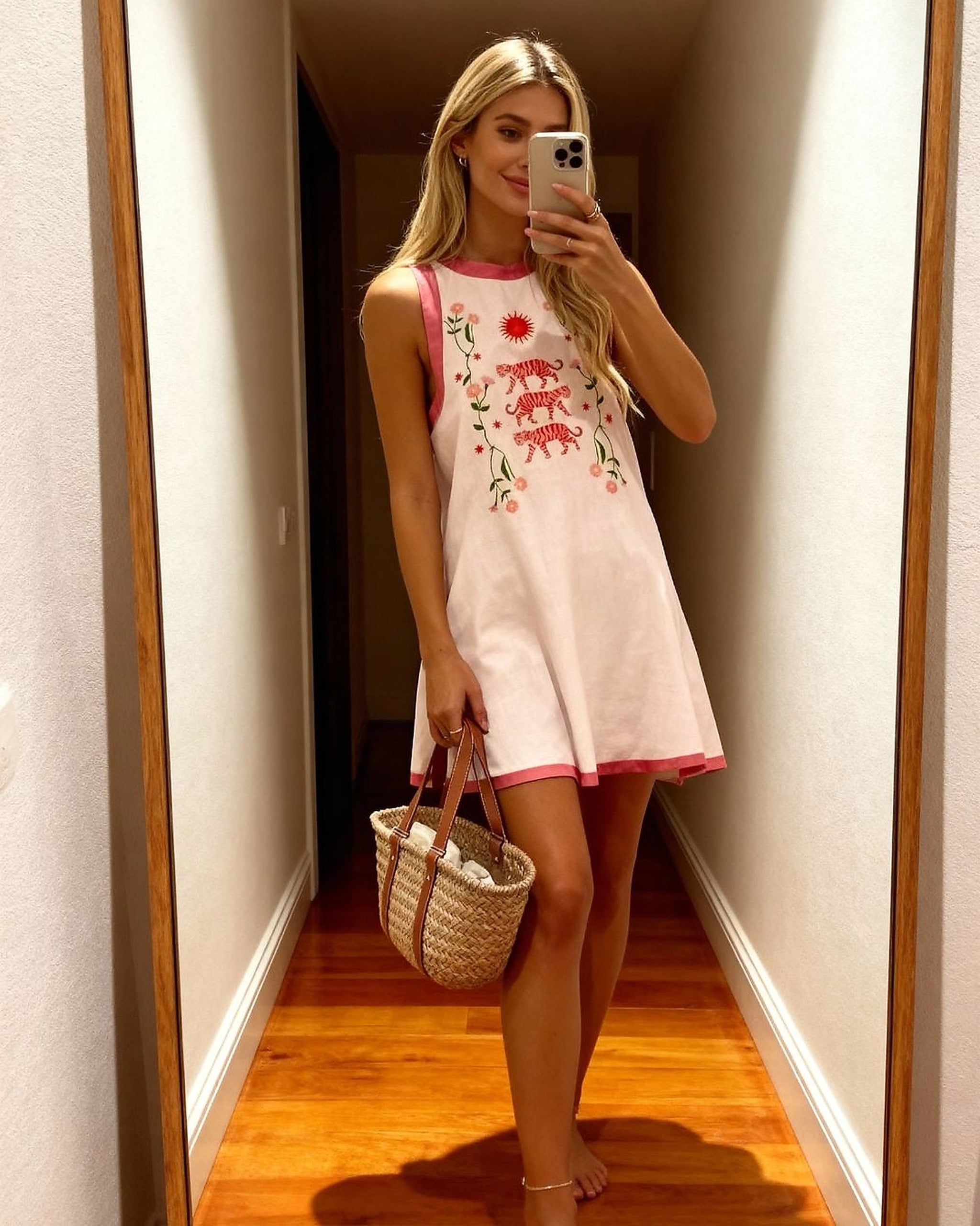 Woman wearing cream and pink Tiger Bloom dress with red floral embroidery, pink trim, holding woven beach bag in mirror