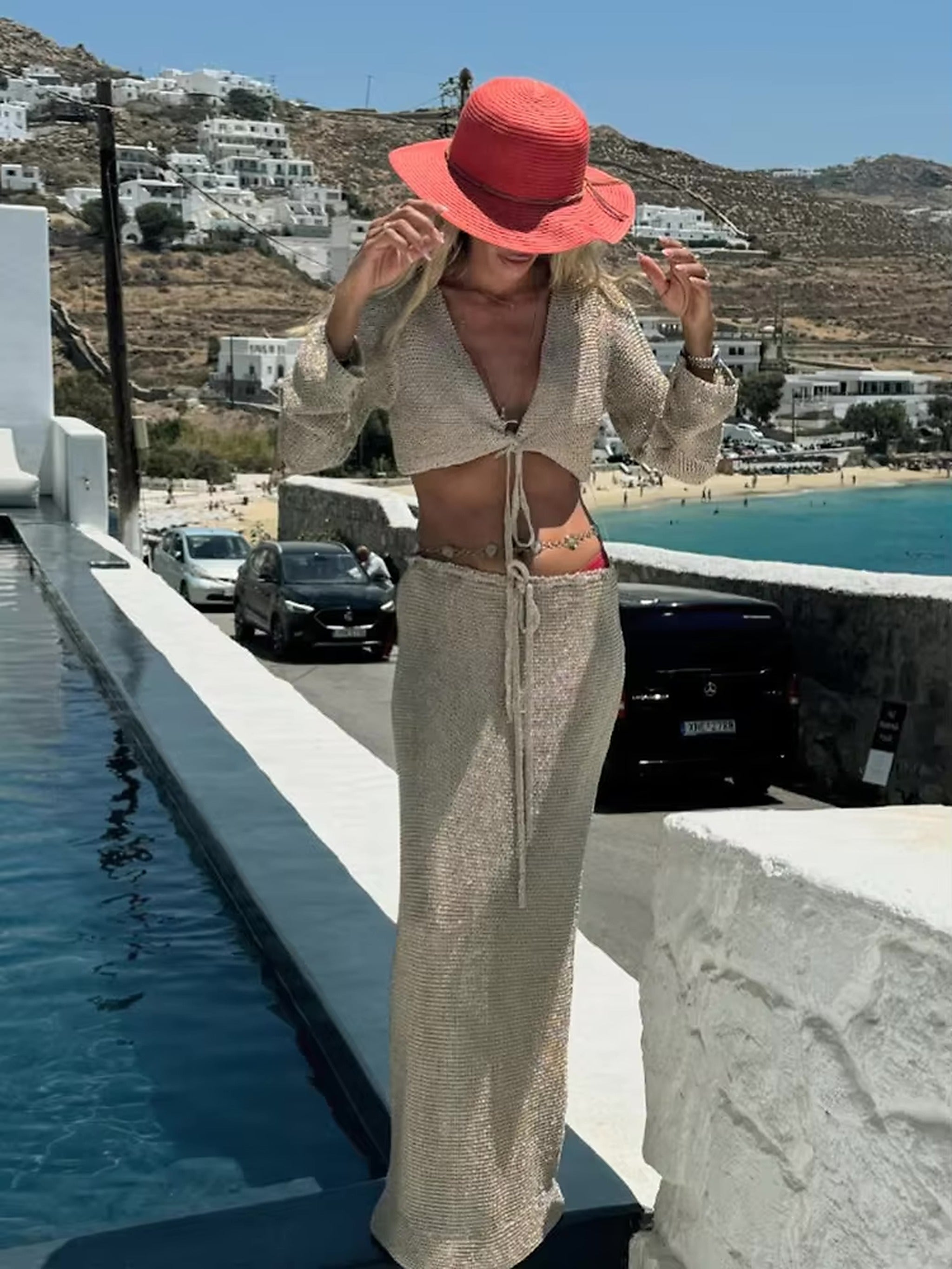 Woman wearing beige knit bikini top and matching sarong with coral sun hat by Mediterranean coastline