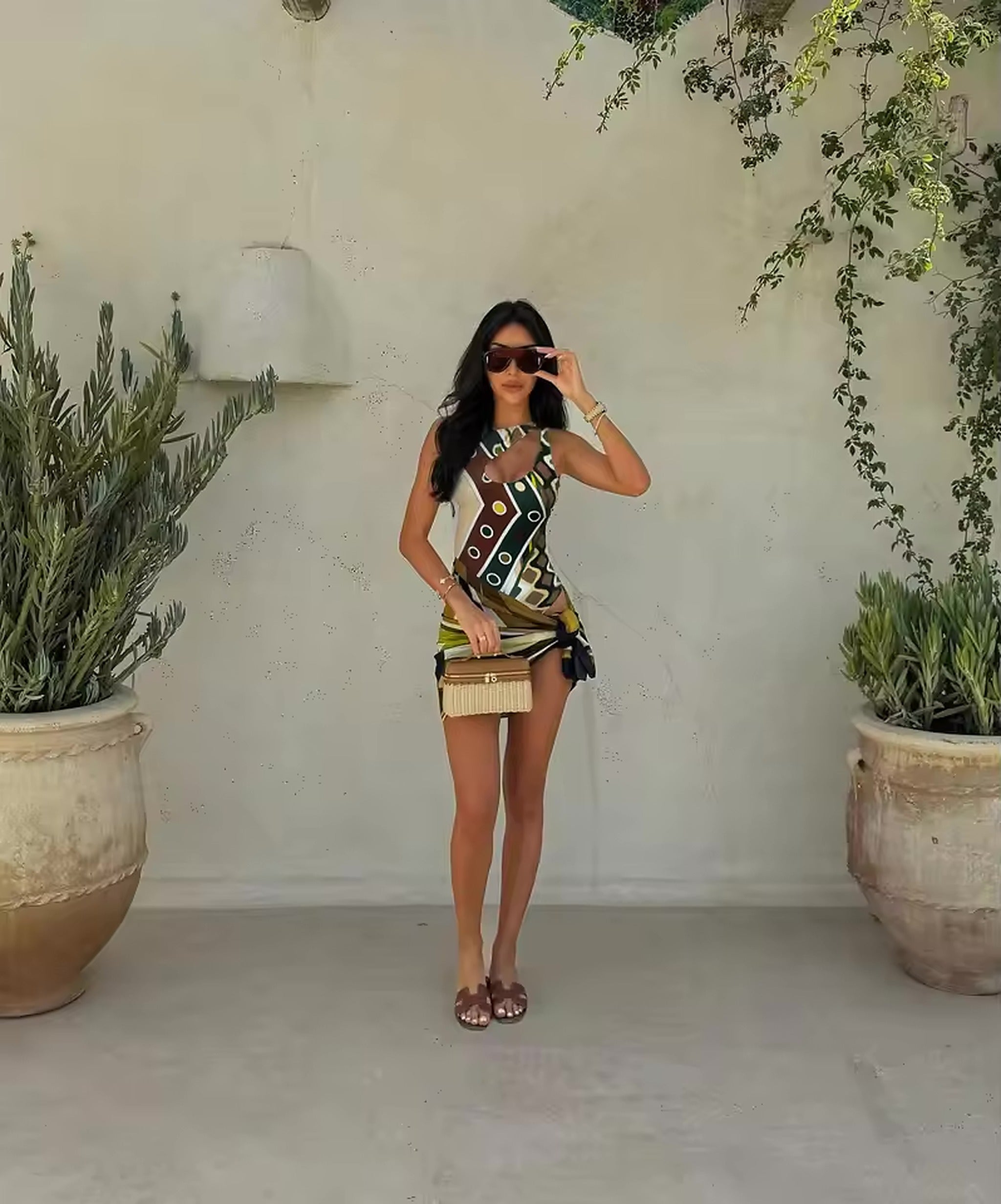 Woman wearing geometric patterned bikini top with fringe skirt, styled with sunglasses and accessories in minimalist garden