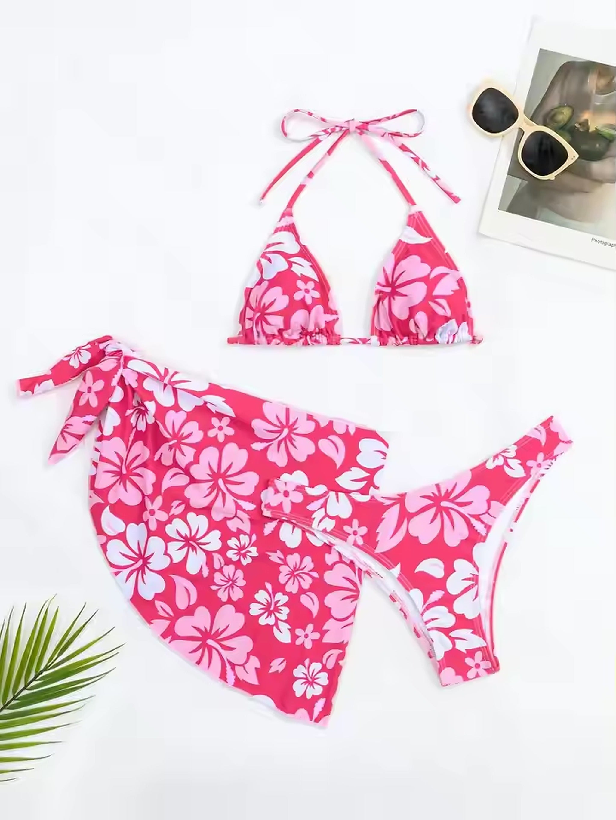 Hot pink floral bikini with halter top and matching swim skirt, styled flat lay with sunglasses and tropical leaves.
