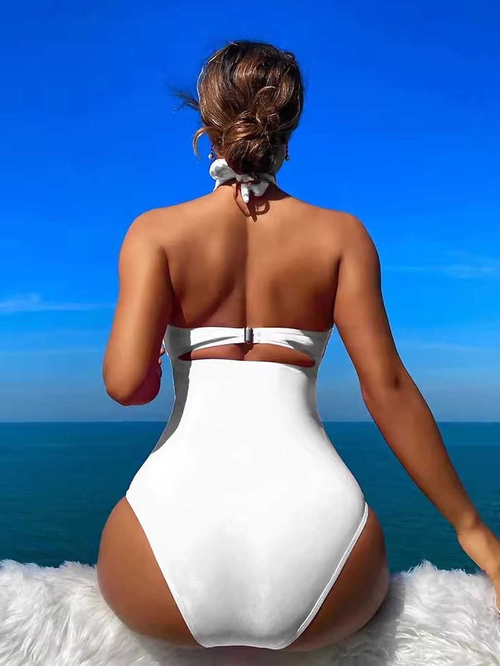 Woman wearing white halter bikini sitting on beach facing ocean under blue sky