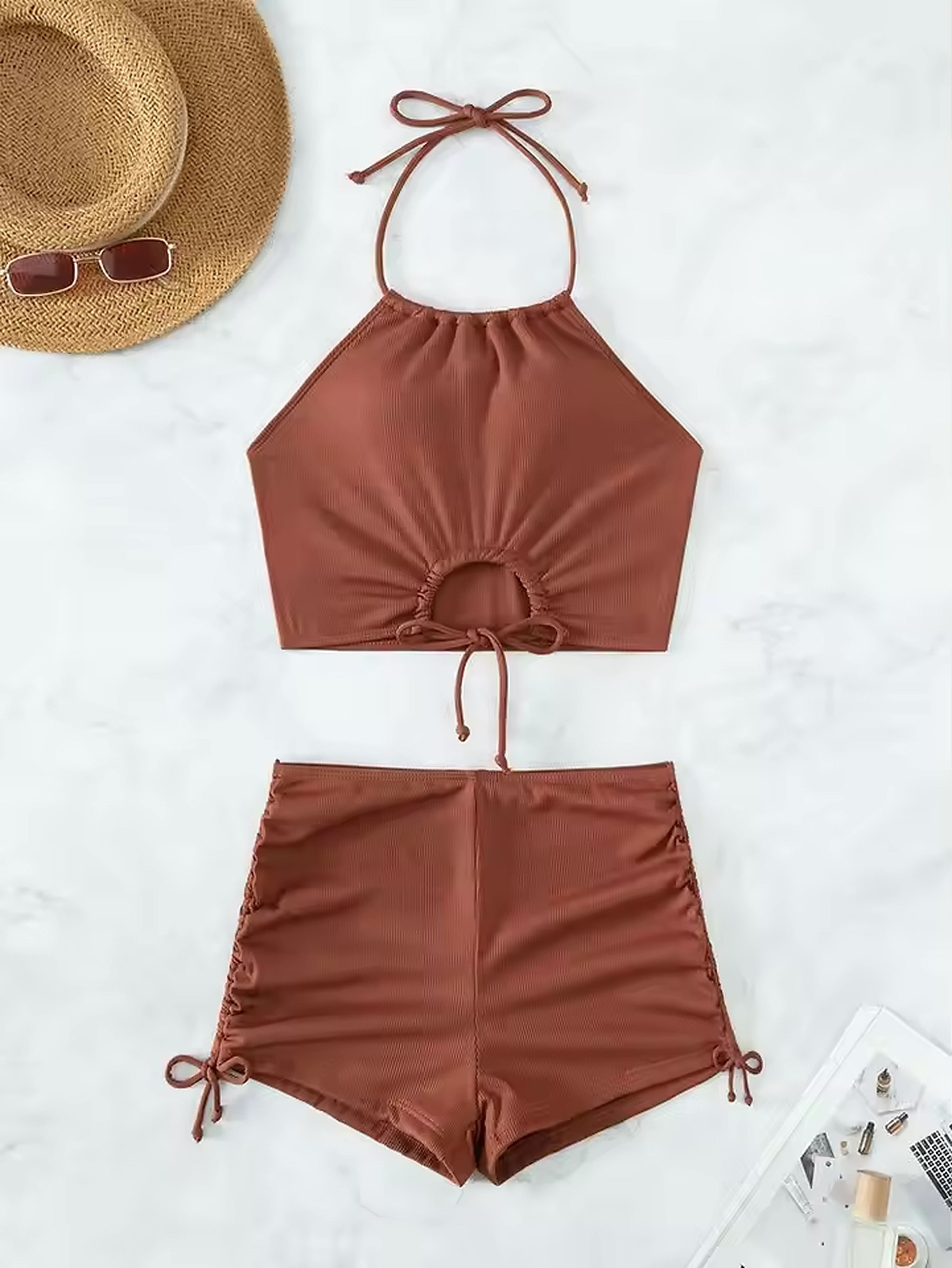 Rust-colored halter bikini set with ruched details and tie-side shorts, styled with straw hat and sunglasses